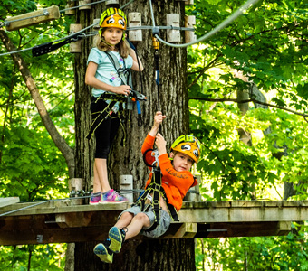 One boy on a zipline while a girl waits on a wooden plank behind him